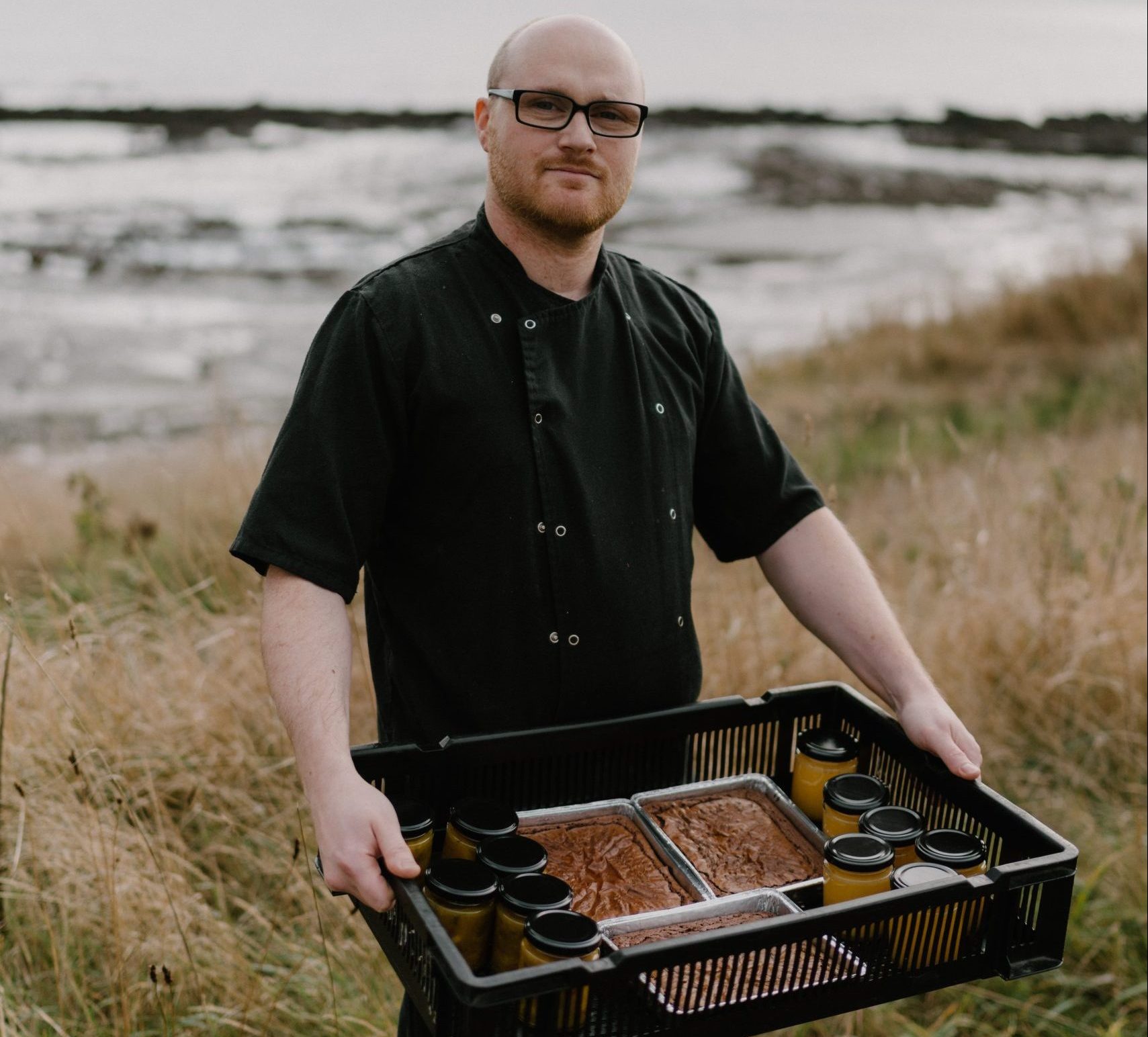 Chef holding condiments and brownies in a tray in front of the sea