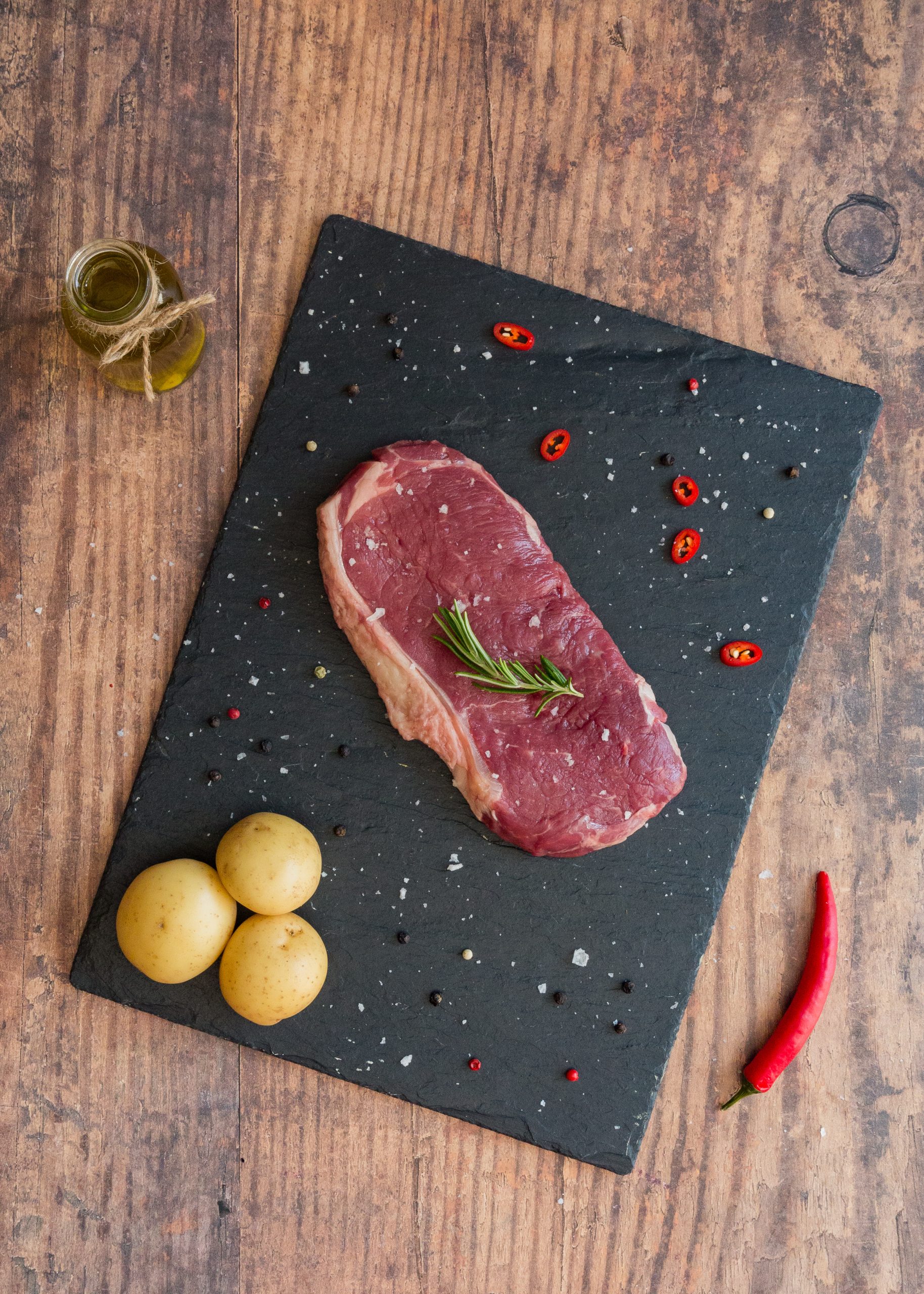 raw sirloin steak on a slate board with new potatoes and a chilli on the side.