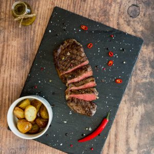 sirloin steak sliced up on a black slate with potatoes in a bowl at the side and a chilli.