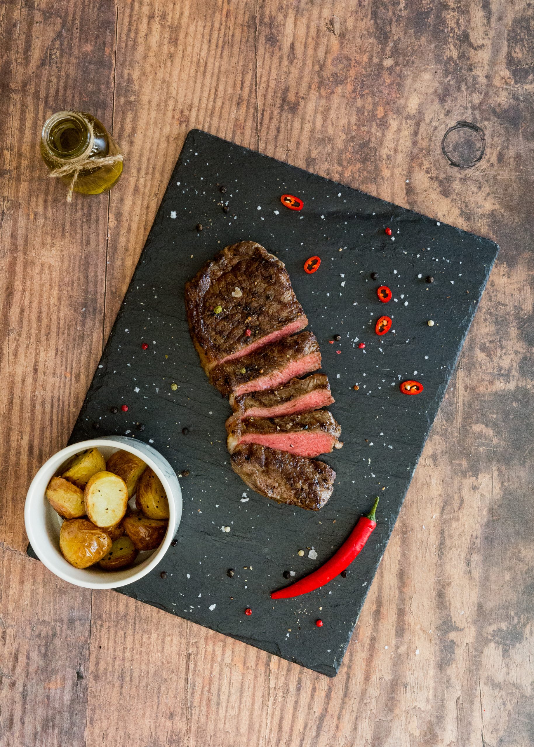 sirloin steak sliced up on a black slate with potatoes in a bowl at the side and a chilli.